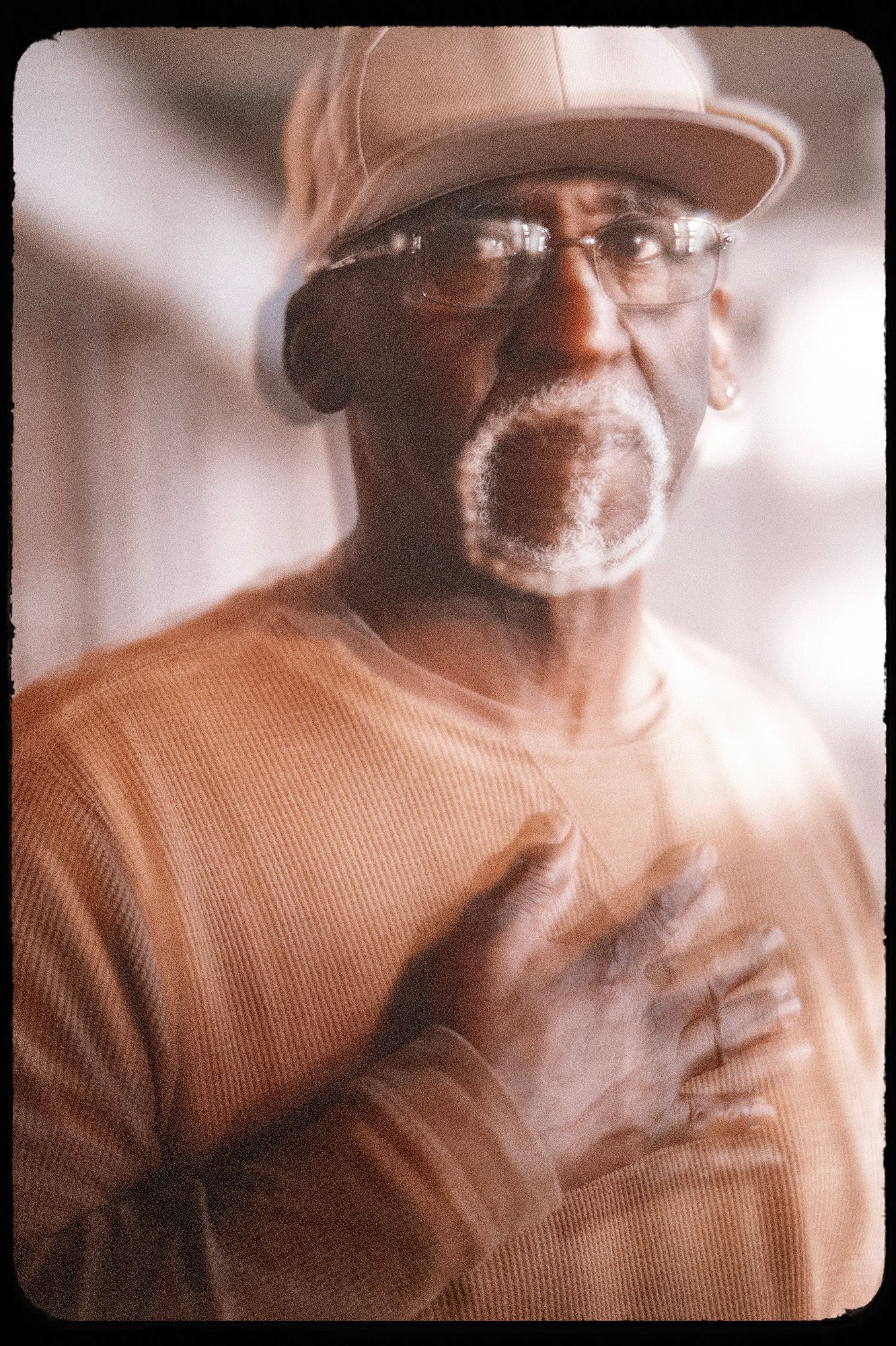 A photo shows Rubin “Jitu” Williams, a Black man wearing glasses, a beige-colored cap and a dark orange long-sleeved shirt, standing with his right palm over his heart. The photo has a shaky effect achieved using a lens. 
