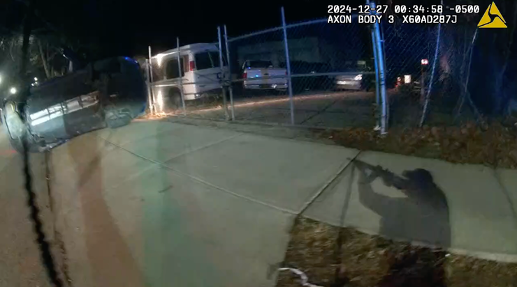 The shadow of a man in a cap holding an assault rifle is visible on the sidewalk at night. A flipped-over car is on the left. Vehicles are parked behind a fence in the background.