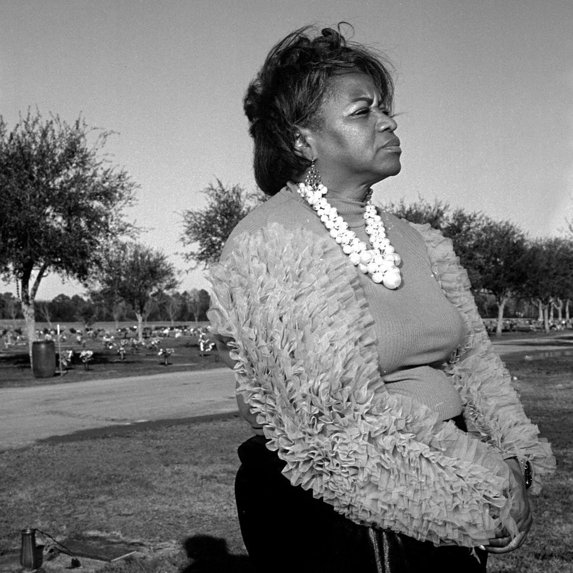A Black woman wearing a white necklace and a shirt with ruffled sleeves stands in a cemetery. There are a few trees behind her. 