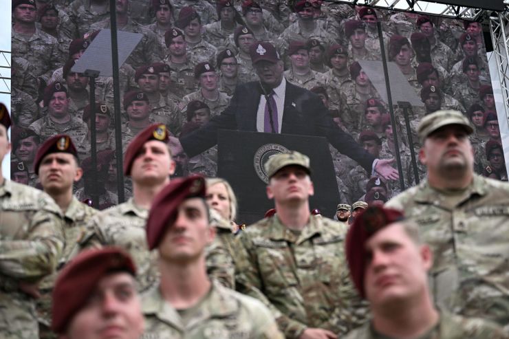 A photo of uniformed soldiers out-of-focus in the foreground, in front of a large screen showing President Donald Trump in a burgundy Fort Bragg baseball cap speaking from a podium with uniformed soldiers standing behind him. 