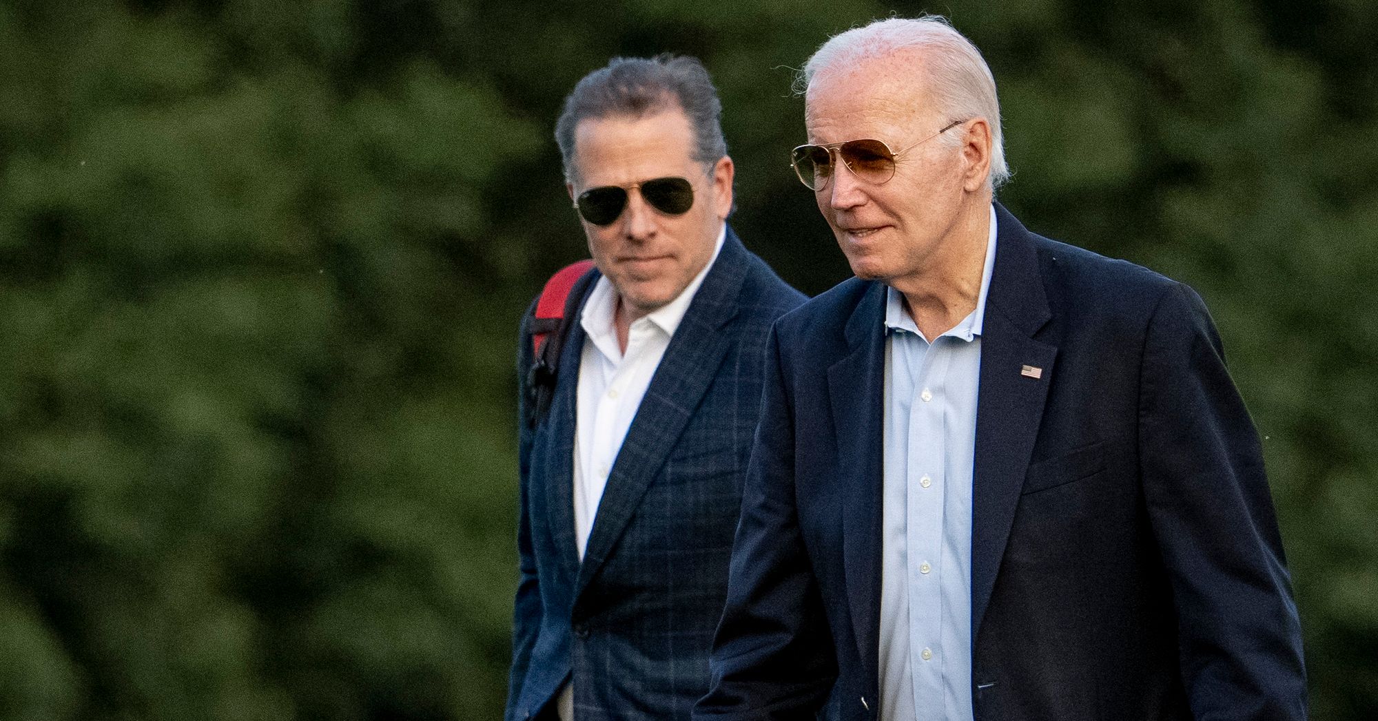 Two White men wearing sunglasses and navy suits are walking in a field. 