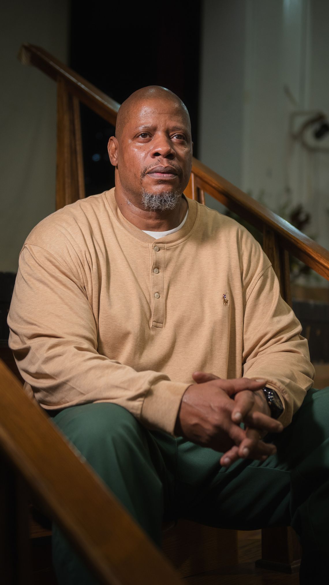 A Black man with a salt-and-pepper goatee, wearing a tan sweatshirt and green prison pants, sits on wooden stairs, his hands folded, as he looks off to the right. 