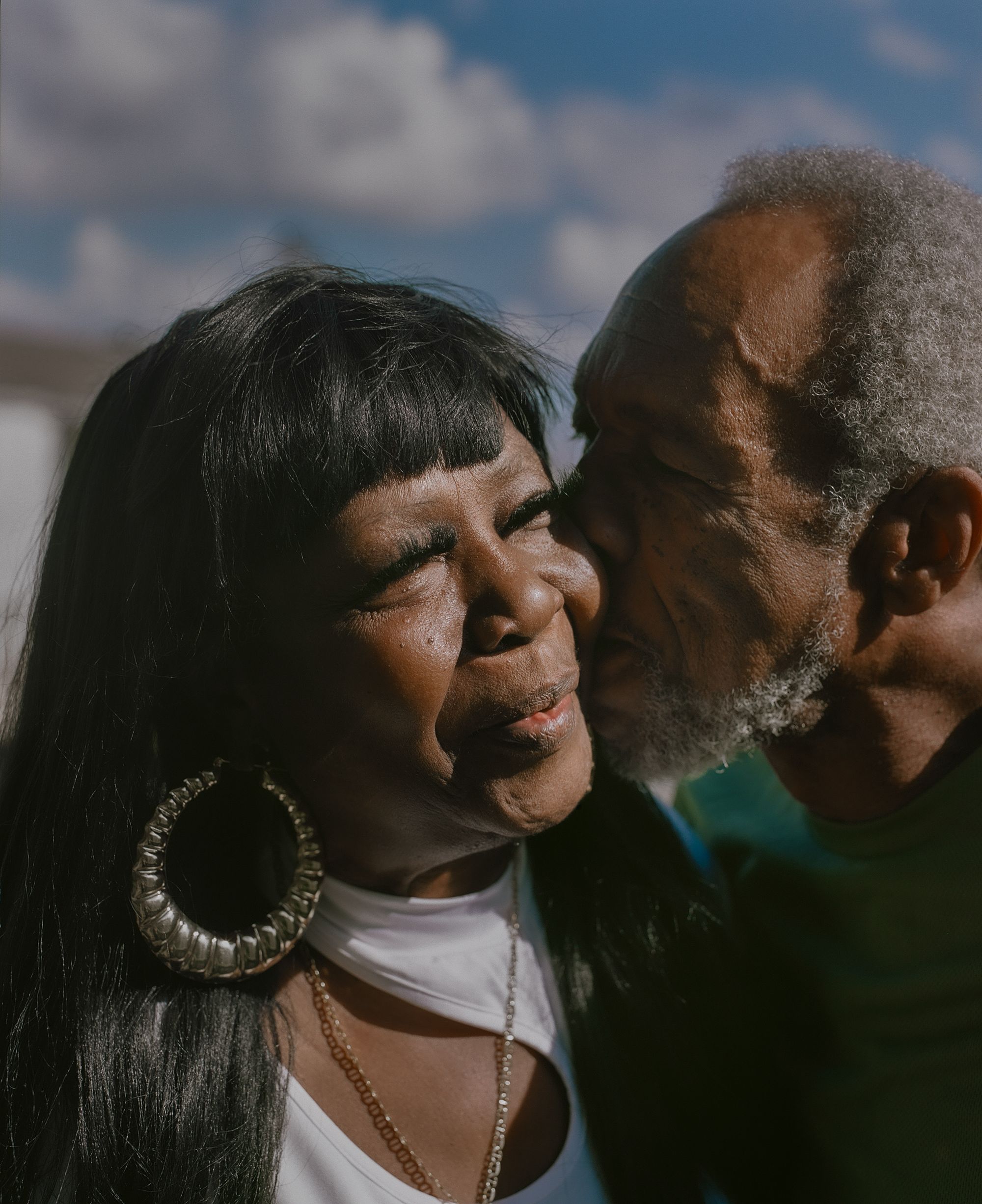 Moses, a Black man, kisses Armour, a Black woman wearing gold hoop earrings, on the cheek.
