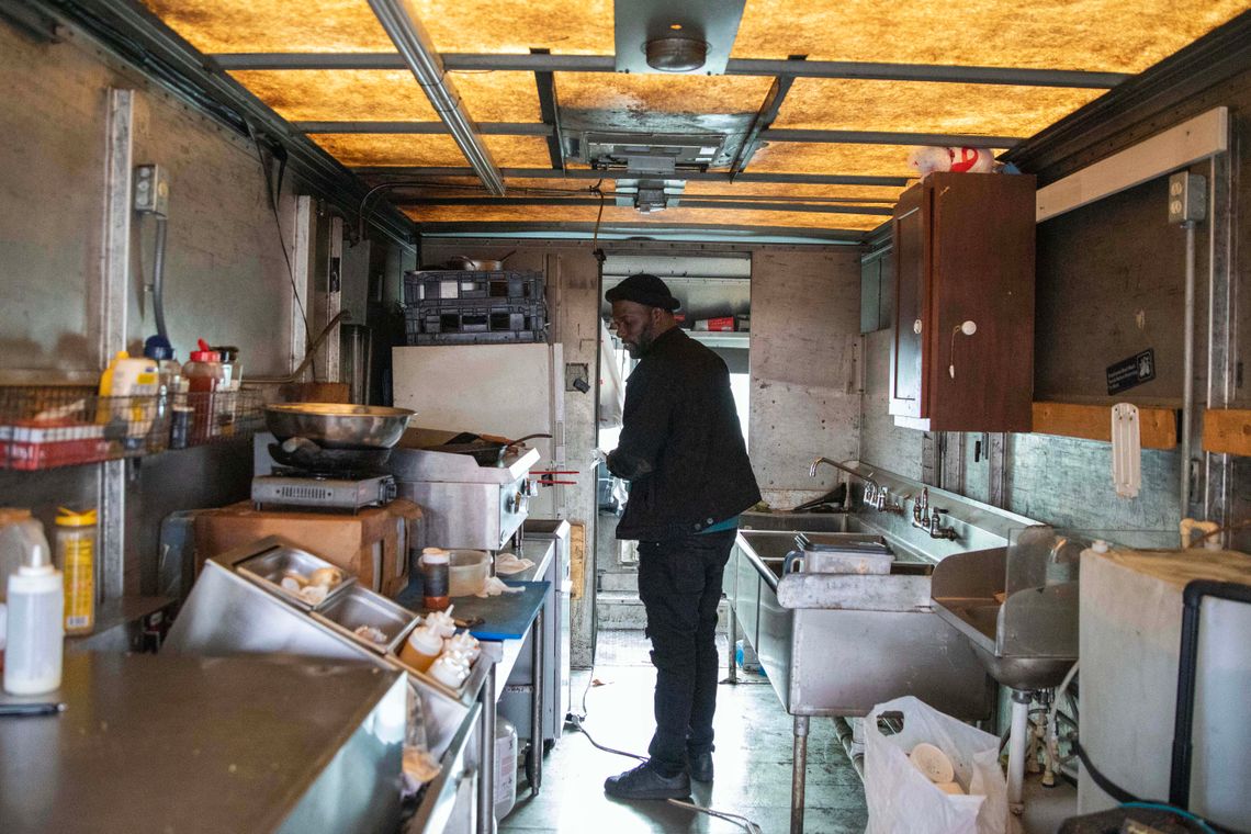 Keedran Franklin, a Black man, stands in the kitchen of his food truck. There are sinks on the right side and a food preparation area on the left. 