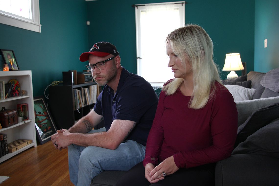 A White man with a beard wears glasses, a red and navy blue cap, a navy shirt and light blue jeans while sitting next to a White woman who is wearing a red long-sleeved shirt. 