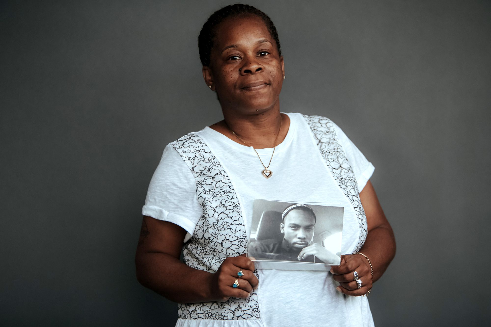 Johndell Gredic, a woman with dark brown skin and wearing a white blouse, stands in front of a gray studio background. She holds a black and white photo of her son Nacear Gredic, who was shot and killed in 2015. 