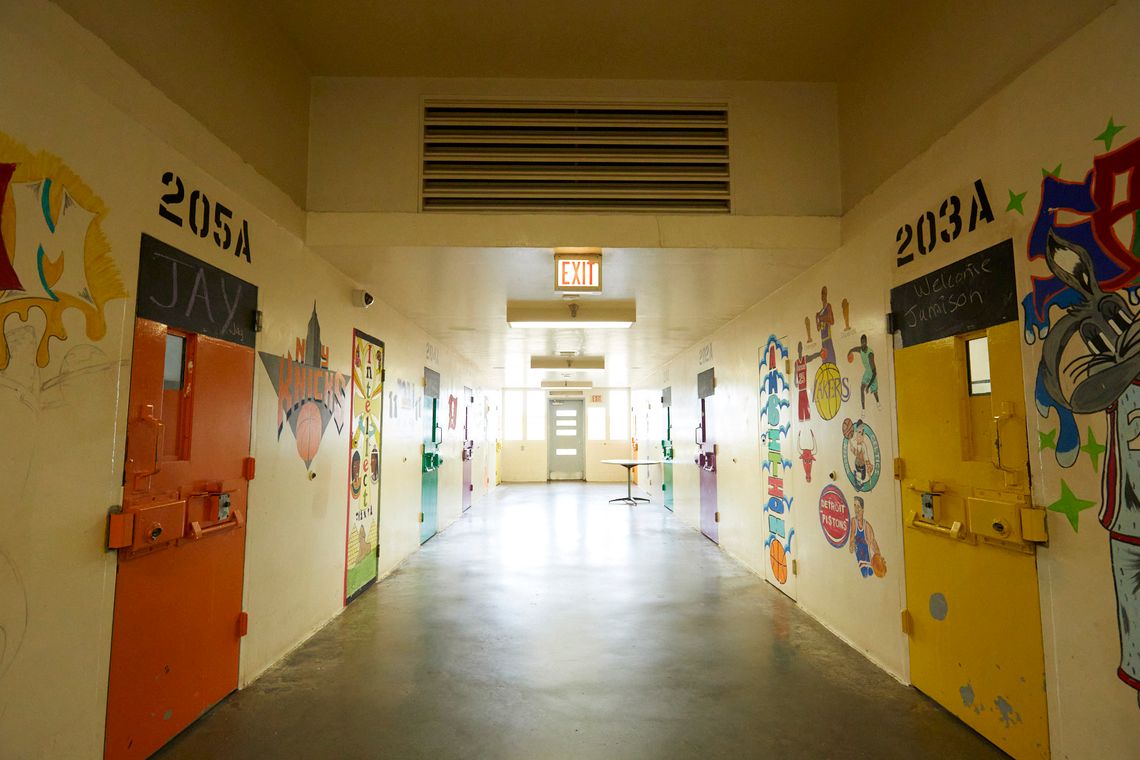 A hallway of the Turbeville Correctional Institution shows the walls covered with colorful logos of basketball teams, including those of the New York Knicks, the Lakers and the Detroit Pistons. On the right wall, the word “Ambition” is spelled out vertically. Above an orange door on the left reads “Jay”, and above a yellow door on the right reads “Welcome Jamison”.