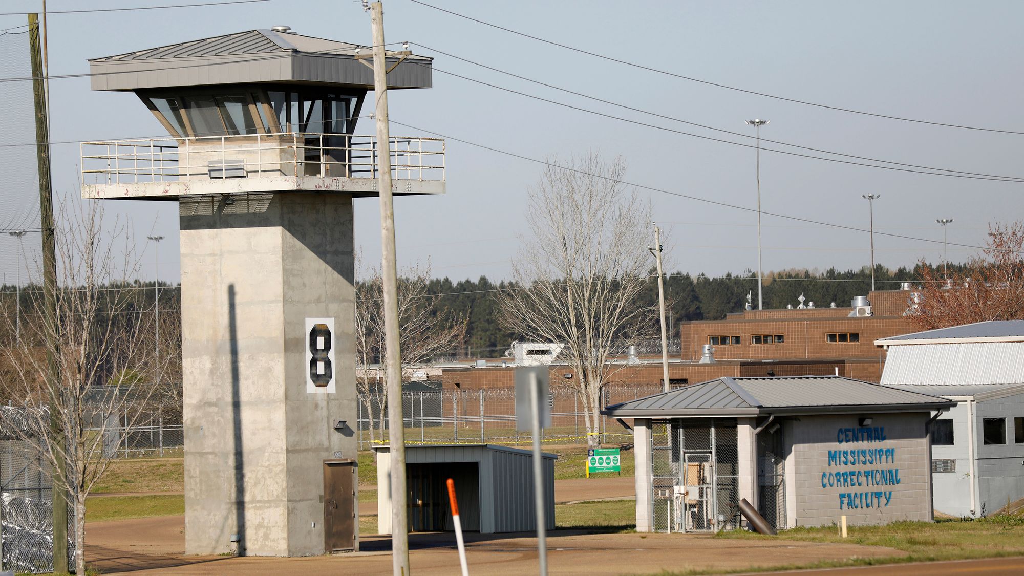 A photo shows a watchtower at the Central Mississippi Correctional Facility. Nearby is a shorter building that has the words "Central Mississippi Correctional Facility" painted on it in blue letters. Behind the watchtower is some fencing and a reddish-orange building.  