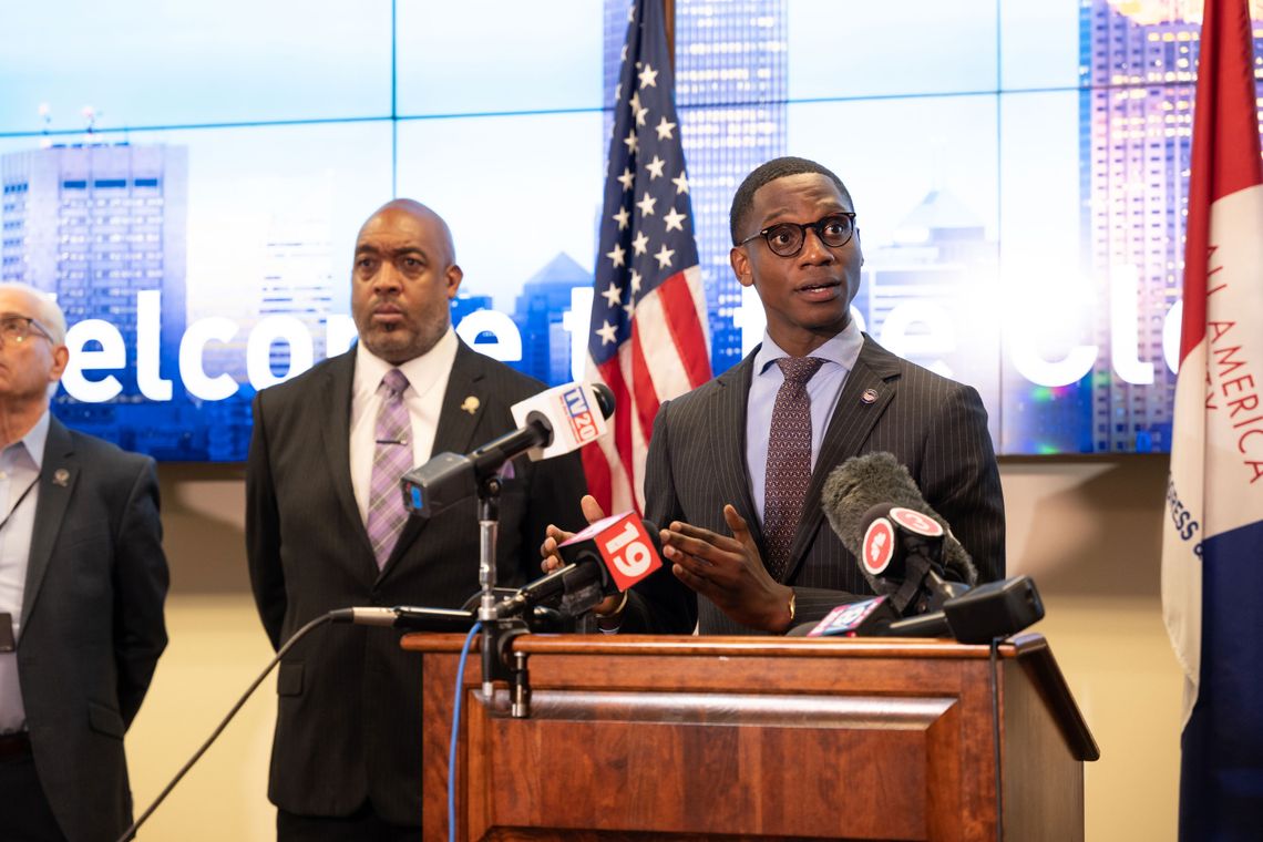 A photograph of a Black man wearing glasses and a suit speaks into news microphones attached to a podium. 