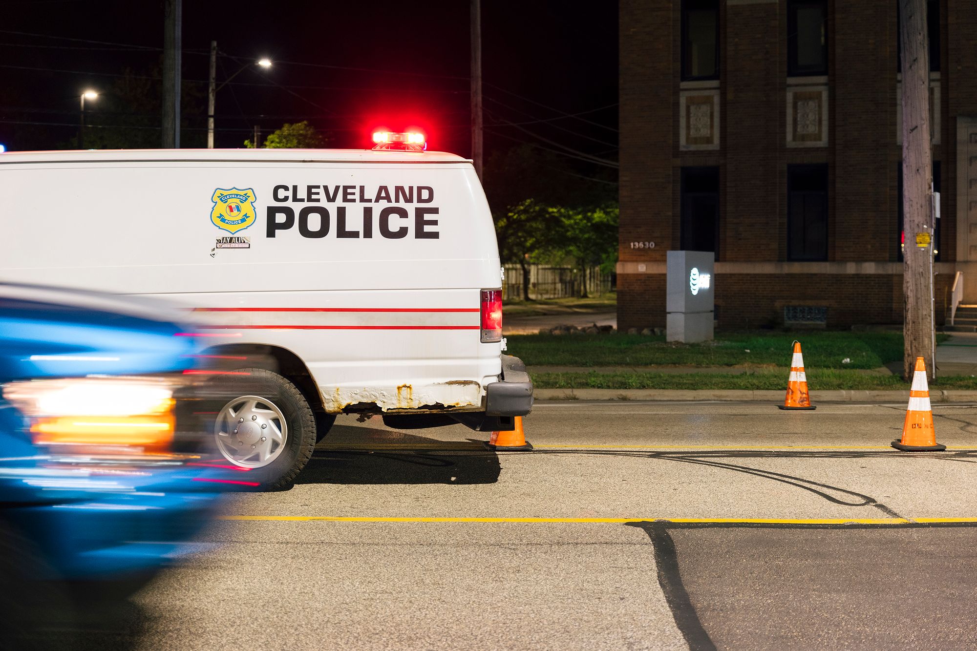 A photo shows a Cleveland Police van on a street at night parked by traffic cones, while a blue car starts to drive by from the left side of the photo.