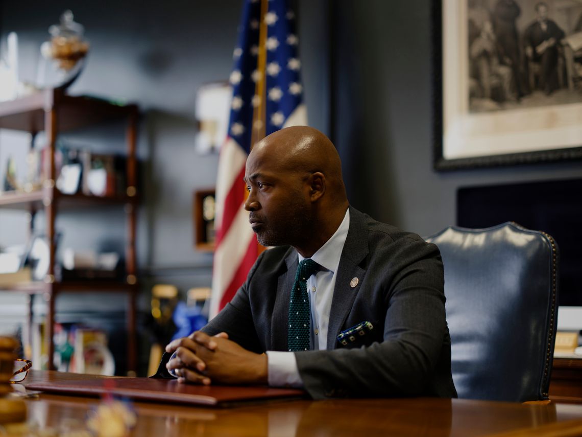 A Black man wearing a gray suit sits at his desk in his office. 