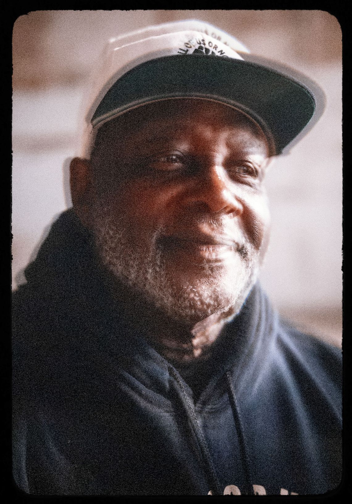 A photo shows a portrait shot of Dorsey Nunn, a Black man with a white beard wearing a baseball cap and a hooded sweatshirt. The photo has a shaky effect achieved using a lens. 