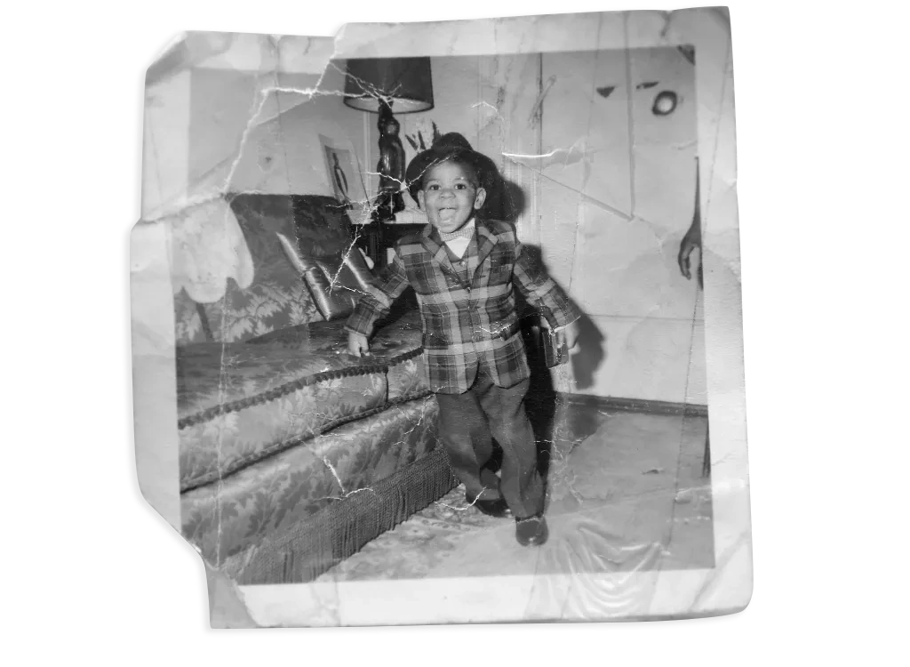 A wrinkled, taped black and white photo shows a smiling Black toddler wearing a fedora, plaid blazer and dress pants, standing next to a couch.