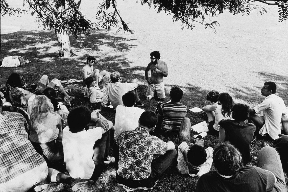 A black-and-white photo shows a bearded White man kneeling and talking to a group of about 20 people, all sitting on the grass in a park.