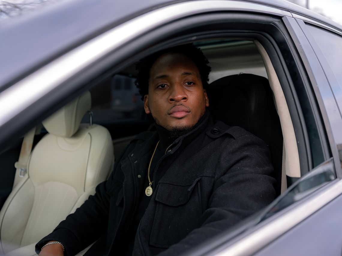 A Black man, in a black turtleneck, black jacket and gold necklace, sits in the driver’s seat of a silver car. 