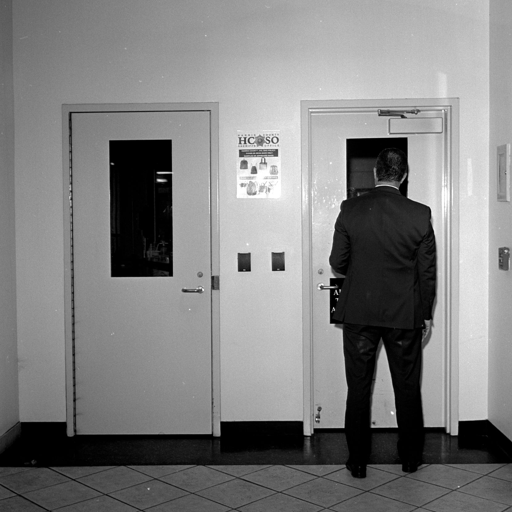 A sign on the wall between two doors has the letters “HCSO” printed on it. A man in a suit, seen from behind, stands in front of the door on the right.