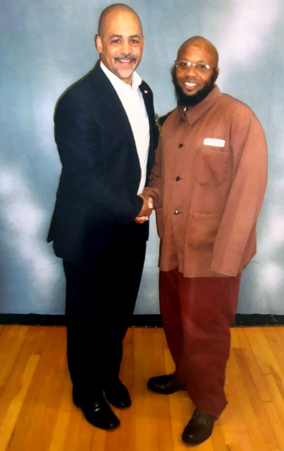 A photo of Tyreem Rivers, right, a Black man with a beard, wearing a brown prison uniform, standing and shaking hands with Rep. Christopher M. Rabb, a Black man, wearing a suit. 