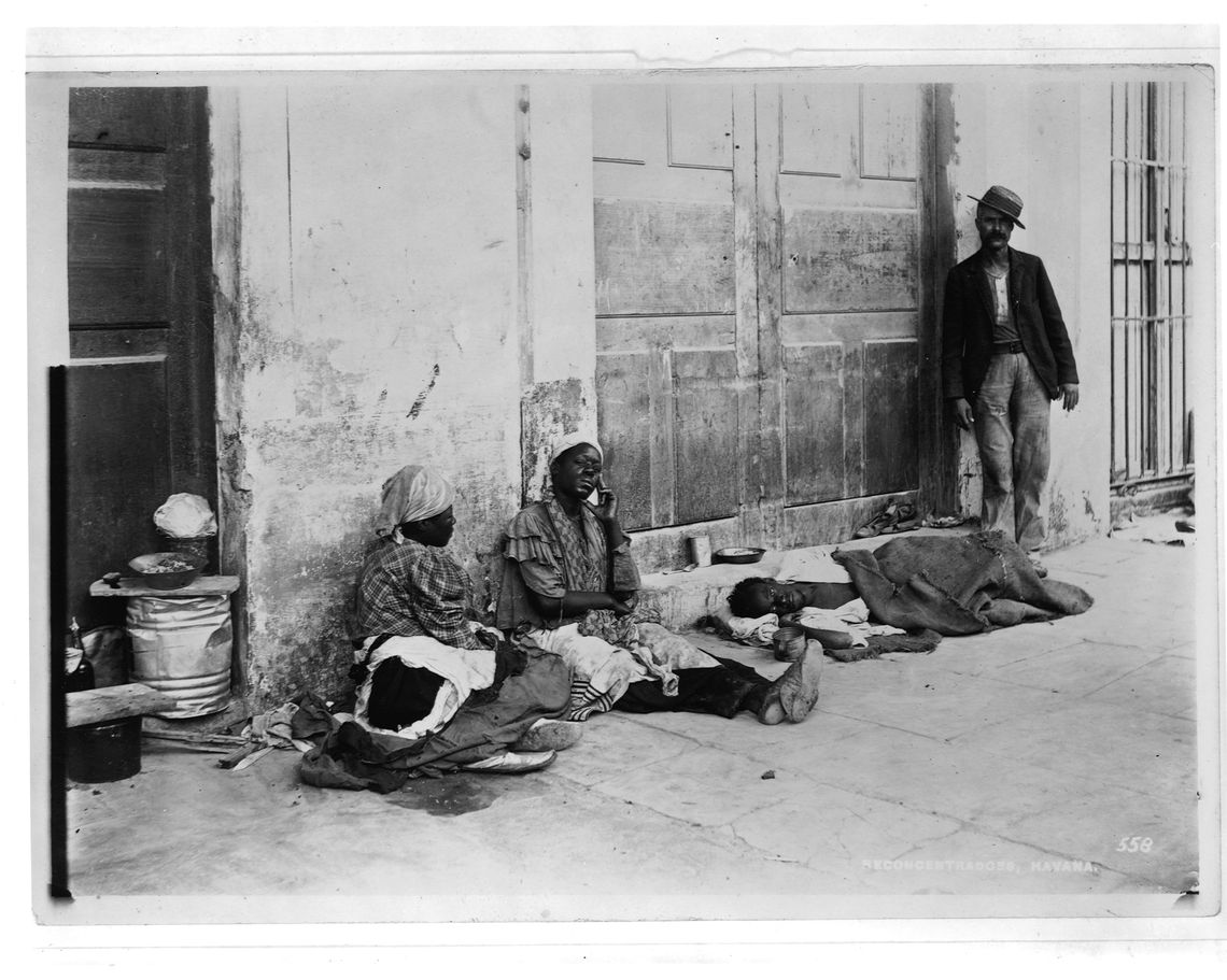 A black-and-white photo shows four refugees sitting on a sidewalk of a concentration camp in Havana in the early 1900s. 