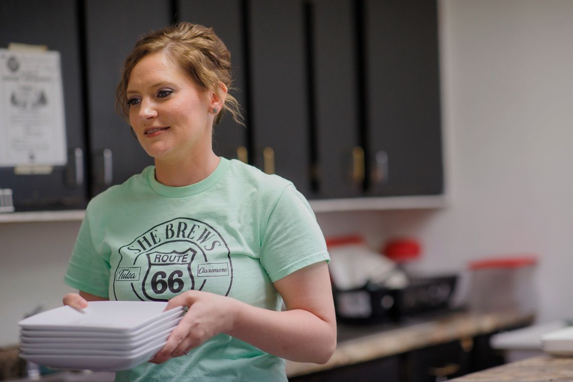 A White woman, wearing a pale green T-Shirt, holds a stack of plates in a kitchen.