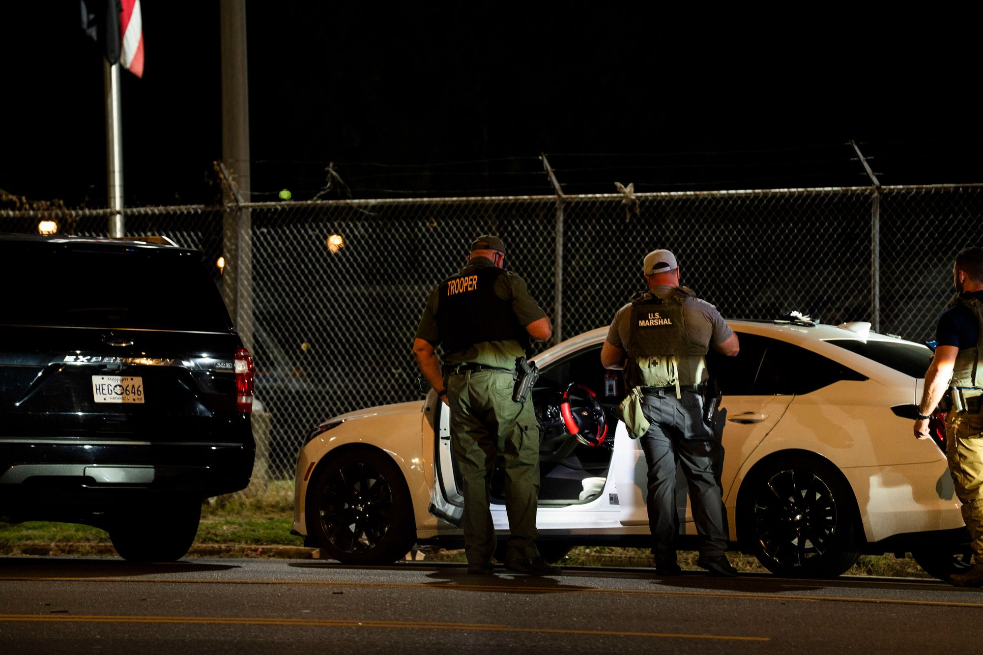 A photo shows officers with vests that read "trooper" and "U.S. Marshal" standing in front of a white car with the door open at night. 