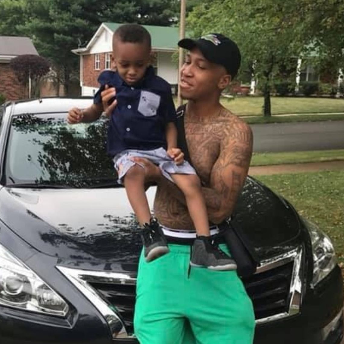 A young Black man with tattoos and without a shirt holds his little son in shorts and a button-down shirt.
