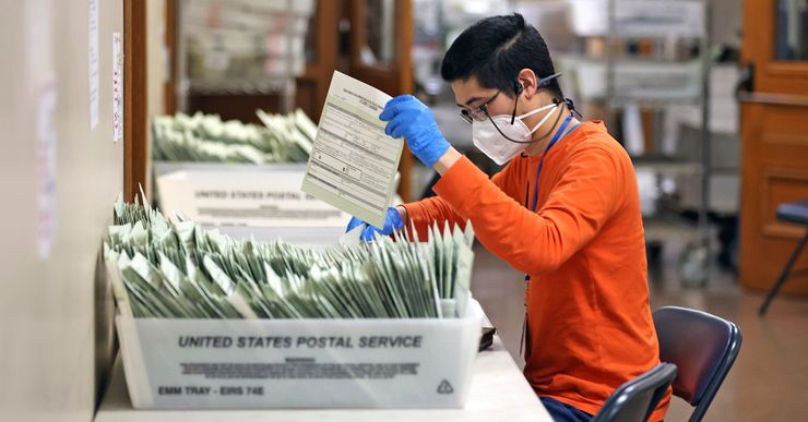 A person with medium-light skin, wearing glasses, a KN95 mask, gloves and an orange long-sleeved shirt, sorts ballots in U.S. Postal Service boxes. 