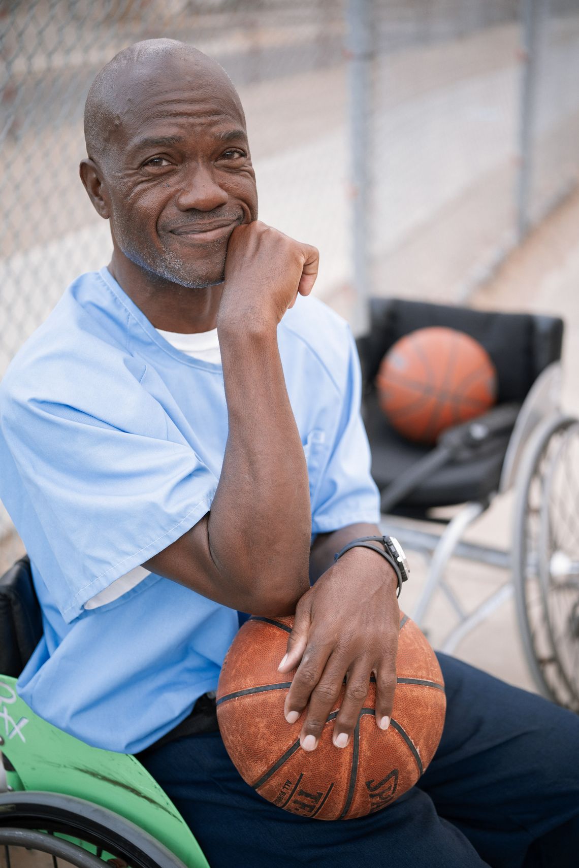 A photo shows a Black man smiling while holding a basketball while sitting outdoors. Behind him is a wheelchair with another basketball in it. 