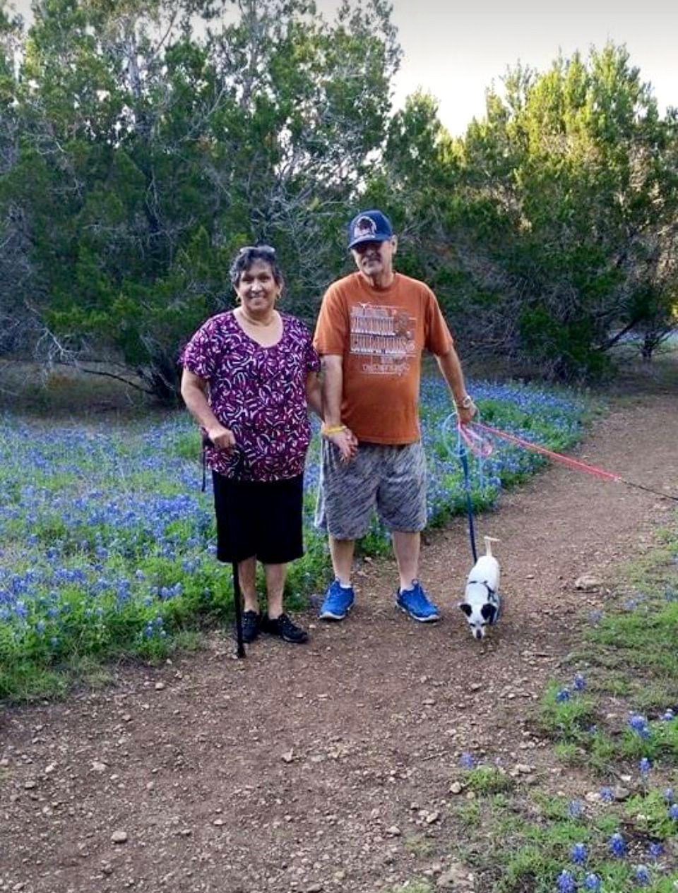 An elderly couple hold hands walking on a pathway in a park. The medium light-skinned woman is also holding a cane in her other hand. The medium light-skinned man holds the leash to a small white dog in his other hand. 