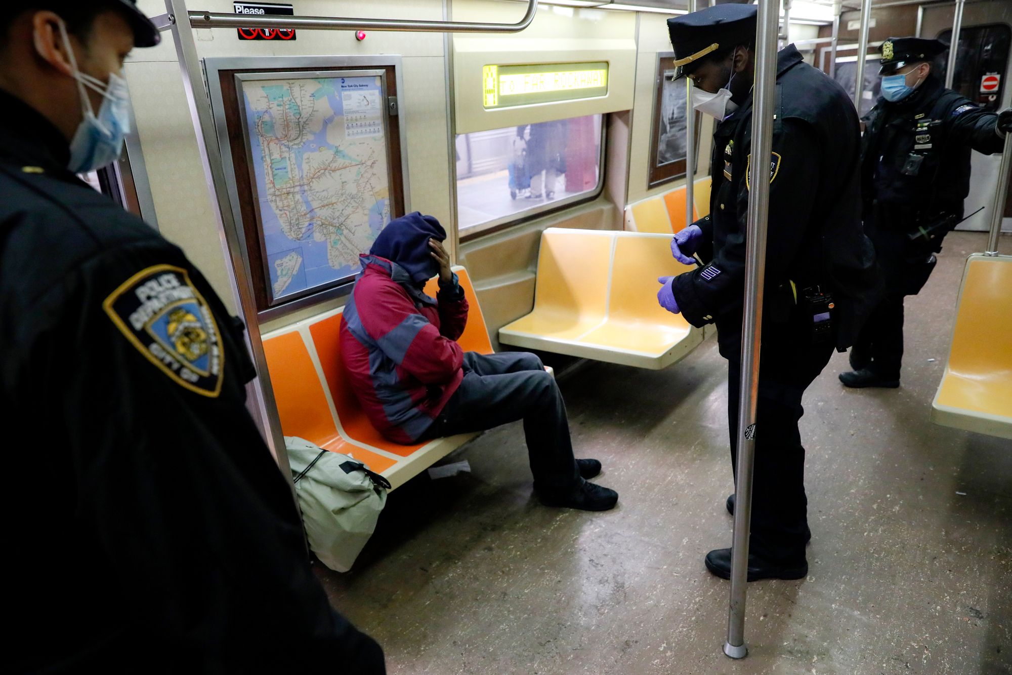 Three standing police officers surround a sitting passenger in a subway car. The passenger sits hunched over, with their hand on their head. 