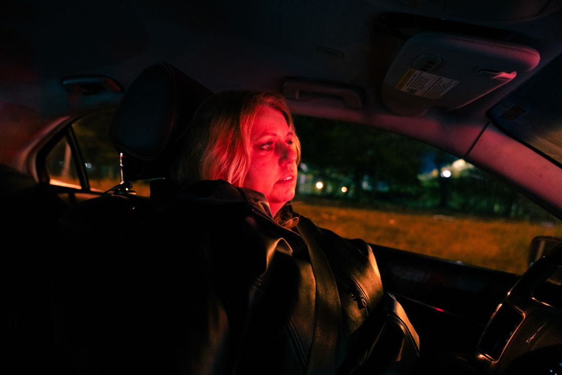 A photo shows a White woman, Sandra Hafraoui, with medium-length blonde hair, sitting in her car at night. A red light shines on her face. 