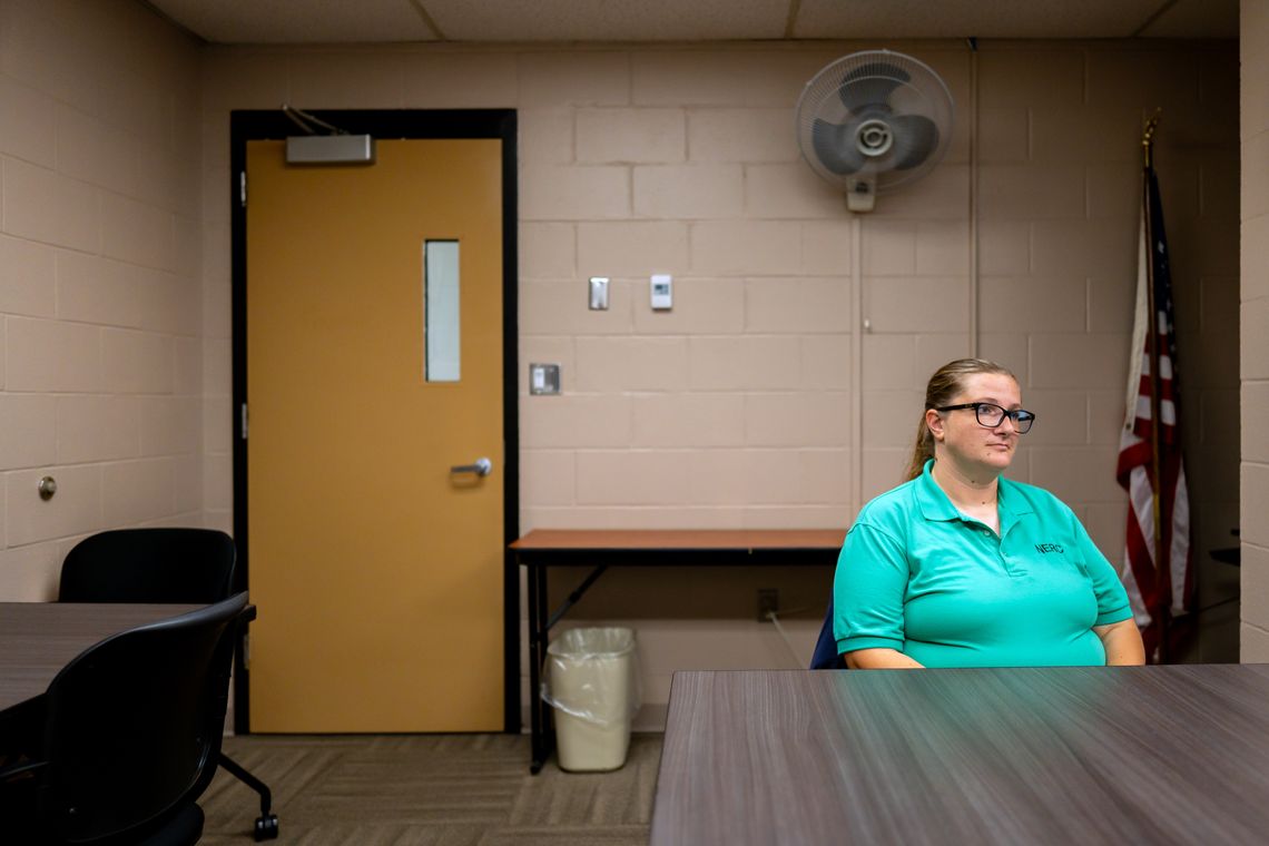 A photo shows Barbara Devine, a White woman with glasses wearing a green T-shirt that is part of a uniform, sitting in a room in front of a table. An American flag is visible behind her on the right, and a yellow door is visible behind her on the left. 