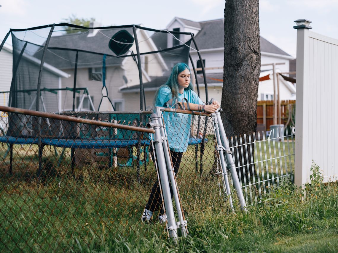 A photo shows a White woman with blue hair leaning against a metal fence. Behind her is a trampoline and a house. 