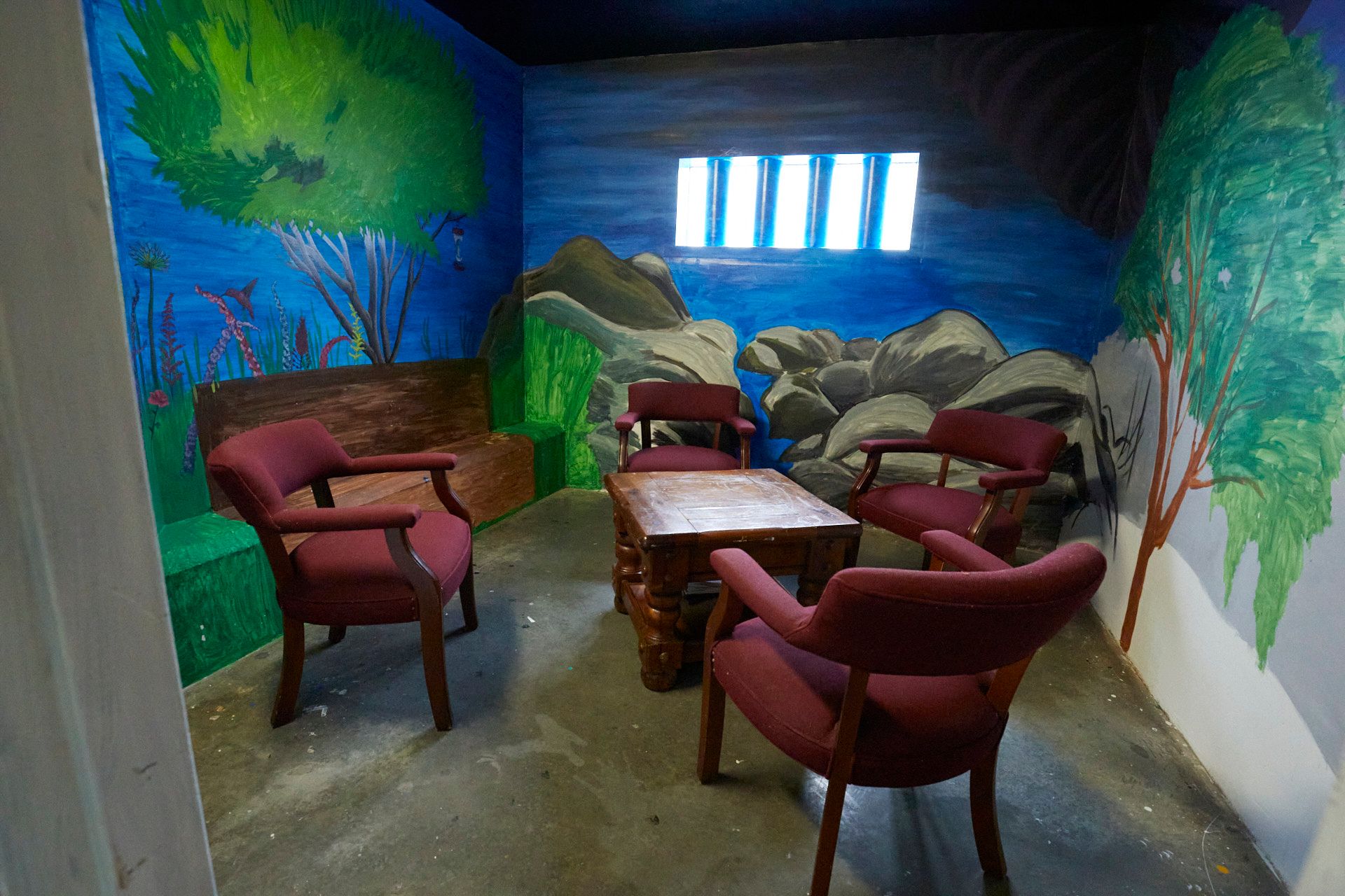 A small room with a window with bars on it has murals on the walls showing trees, rocks and plants. Four cushioned, burgundy chairs sit around a small wooden coffee table.