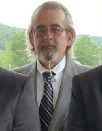 A White man with white hair wears a gray suit jacket and a striped navy blue tie. Trees appear in the background. 