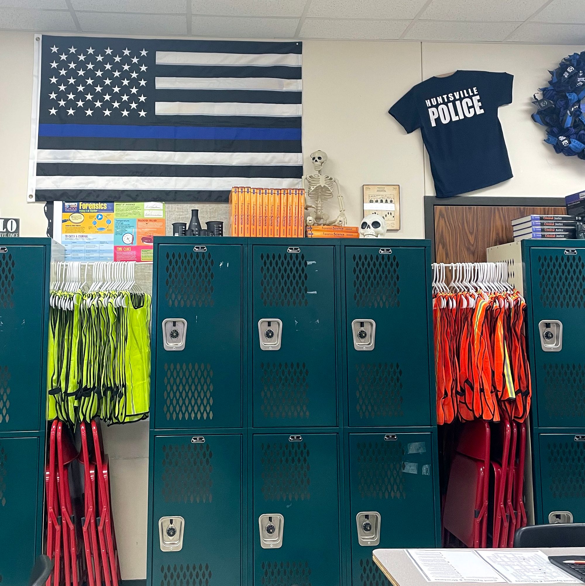 A locker room with green lockers, fluorescent orange yellow safety vests hanging on a rack. On the wall is a black and white U.S. flag with a dark blue line replacing a white strip and a T-shirt that reads, “Huntsville police.” 