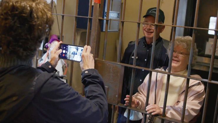 Two older White tourists stand behind the bars of a jail cell to be photographed by a woman holding a camera phone.