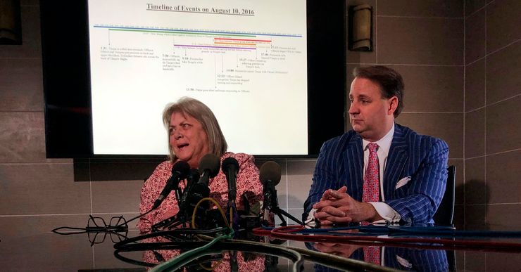 A White woman, wearing a red and pink patterned shirt, speaks into several news microphones inside a conference room. A male lawyer, in a red tie and blue pinstriped suit, sits next to her with his hands clasped on the table. 
