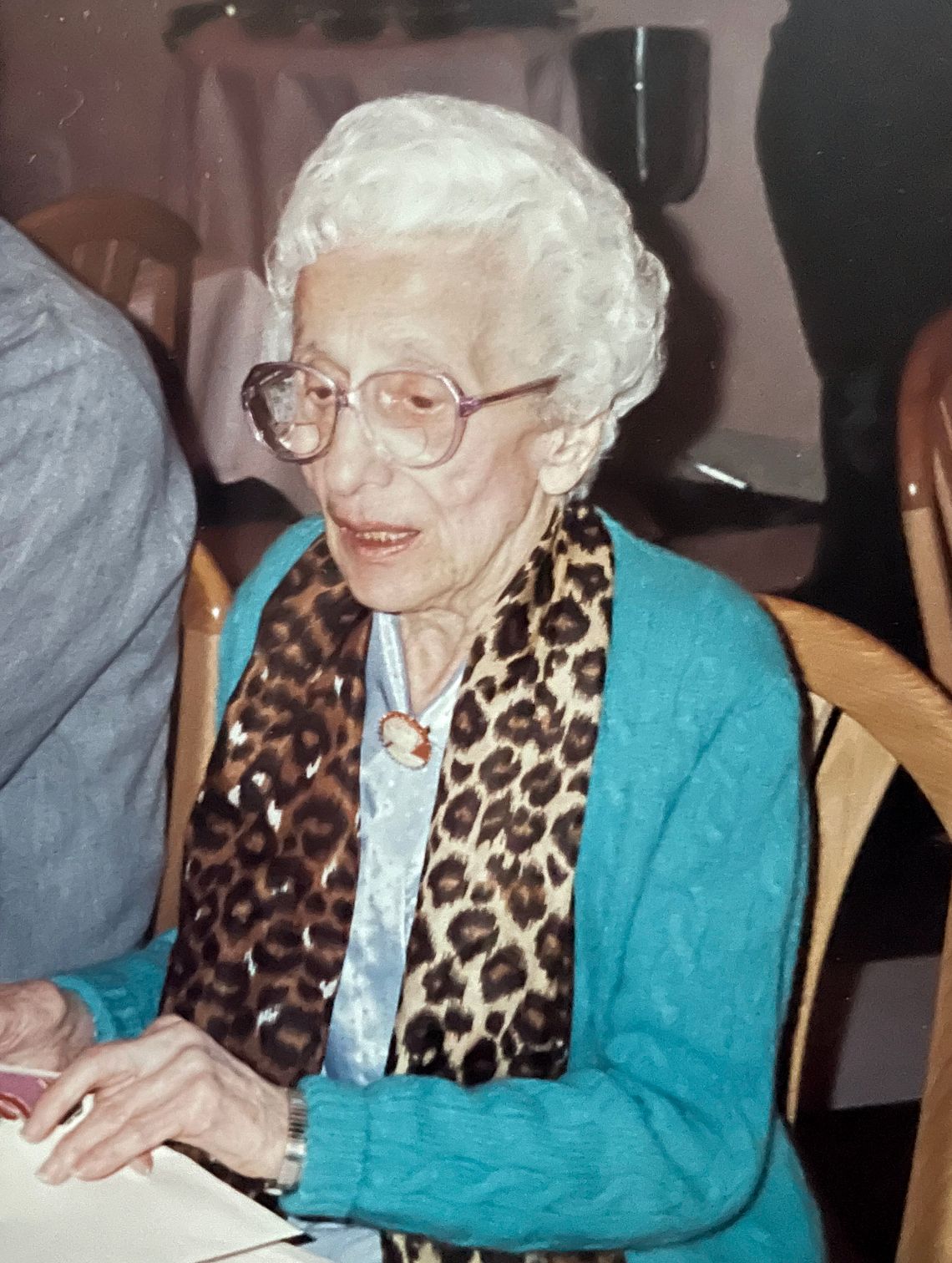 A photo of Mary Tonzola, an elderly White woman with short white hair and glasses, wearing a teal cardigan and sitting at a table. 