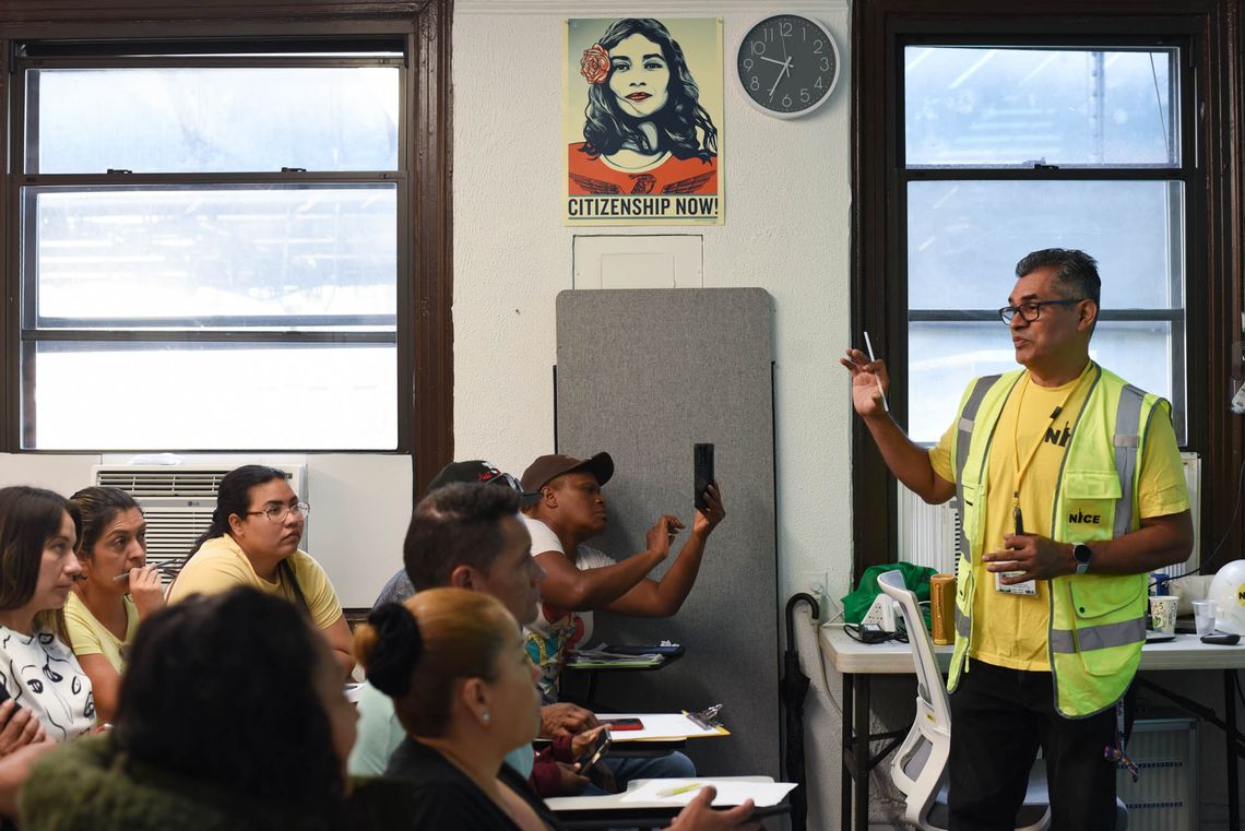 A group of people representing a mix of genders and ethnicities sit in a classroom at school desks. At the front of the room is a man in a yellow shirt wearing a reflective vest that says “NICE.” He has one hand up as he speaks. In the background, on the wall of the classroom, is a poster that reads “Citizenship Now.”