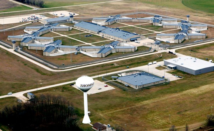 An aerial view of a U.S. penitentiary in Thomson, Illinois. A white water tower, with the word "Thomson" painted on it is in the foreground, next to a grove of trees. 