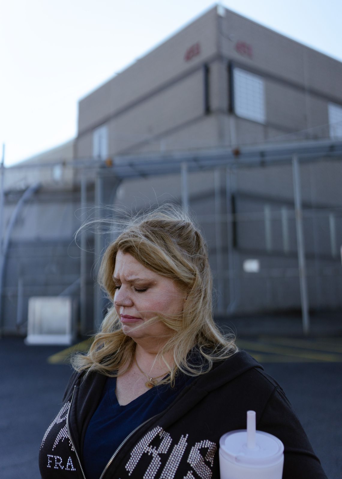 A photo shows a White woman, Sandra Hafraoui, with medium-length blonde hair, standing outside Delaney Hall, an ICE detention center. She is wearing a black, zip-up hoodie sweatshirt, and metal fencing and a two-story building is visible behind her. 