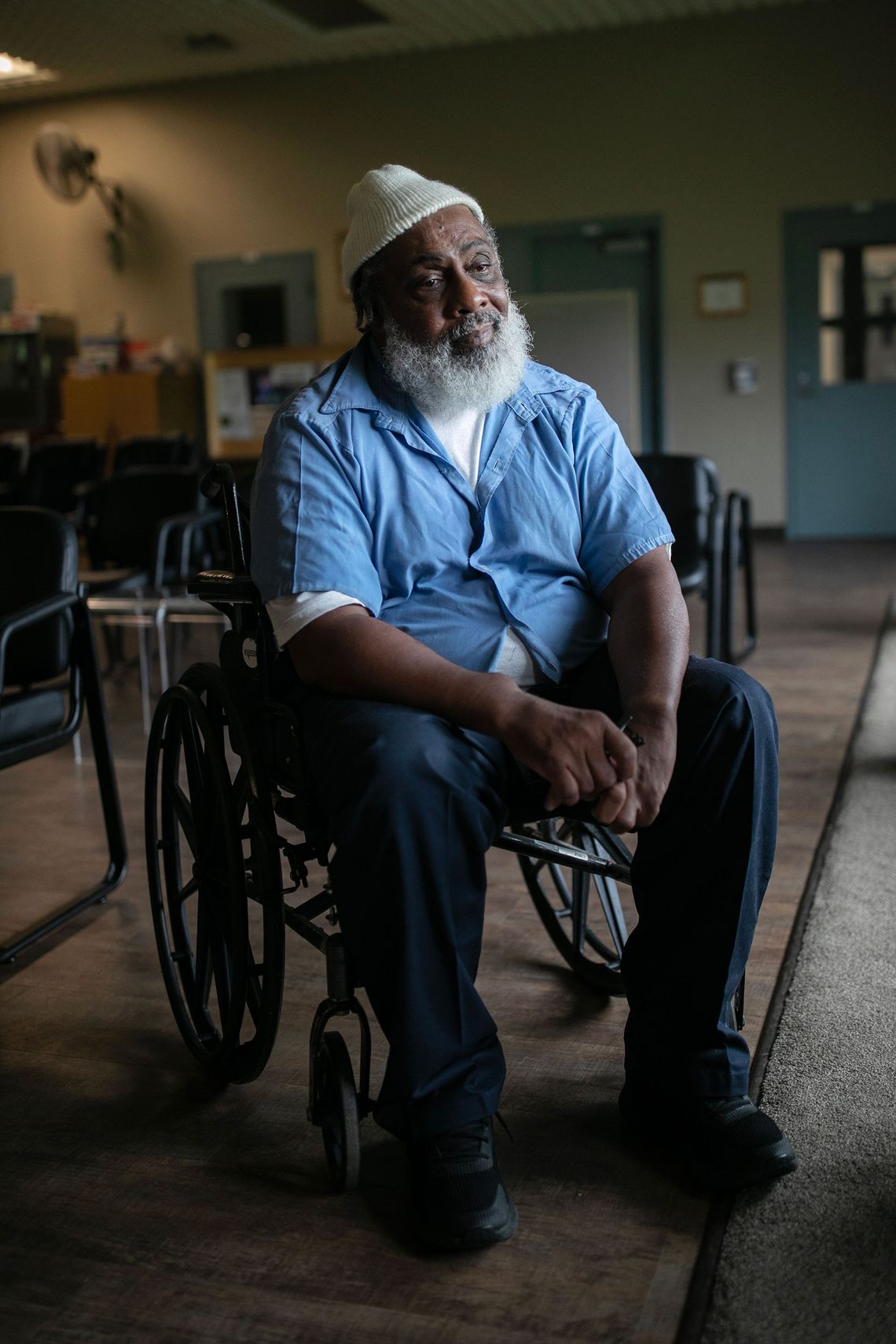 An older Black man — a wheelchair user with a gray beard, white knit hat and blue shirt — looks off-camera. 