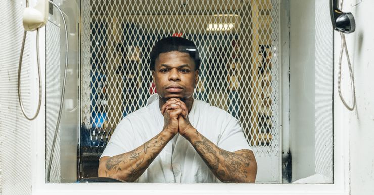 Terence Andrus, wearing a white prison uniform, looks through glass.  A phone hangs on either side of the divider.  