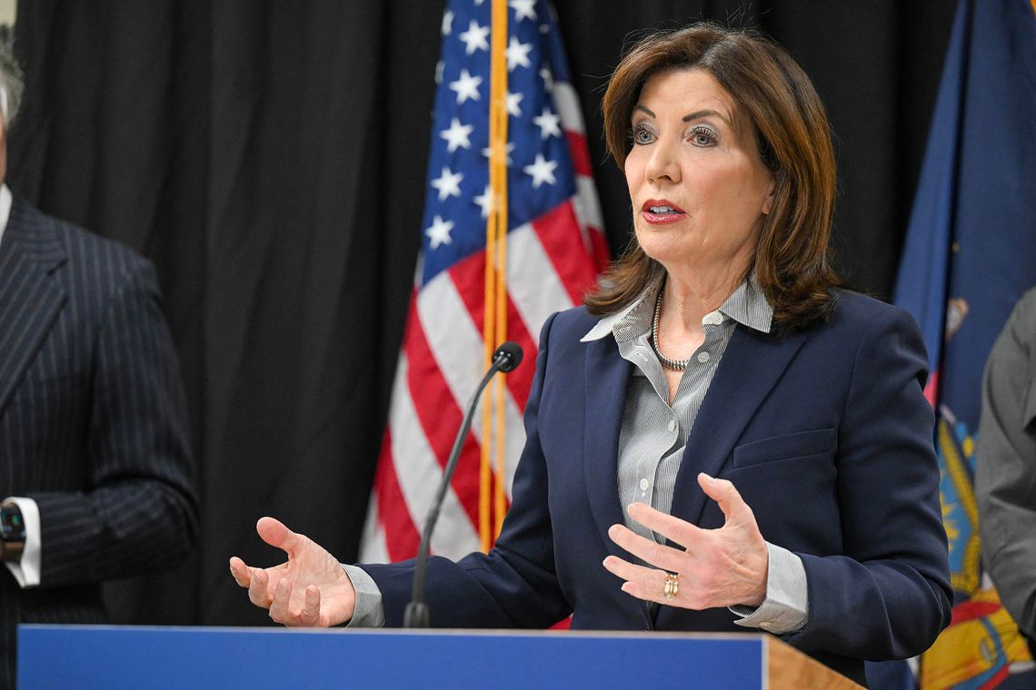 A photo shows a White woman wearing a navy blazer and a gray blouse speaking into a mic while standing at a podium. A U.S. flag is in the background. 