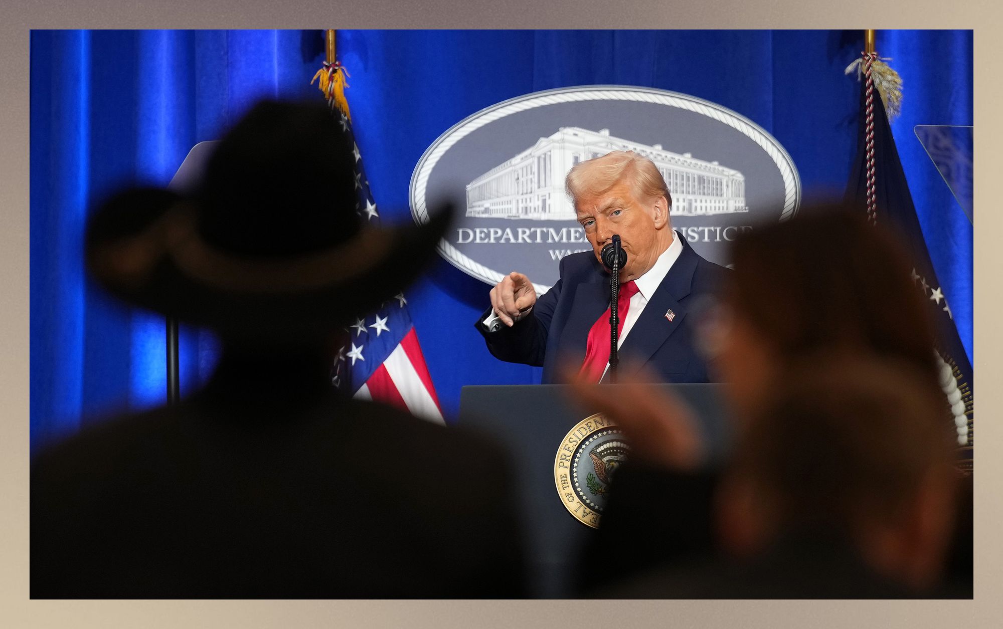 President Trump, a White man wearing a navy blue suit jacket, white button-down shirt and red tie, points at members of the audience. In the foreground of the photo, there are silhouettes of audience members, including one person wearing a cowboy hat. Behind Trump is a cobalt blue backdrop and a sign that says “Department of Justice.” 