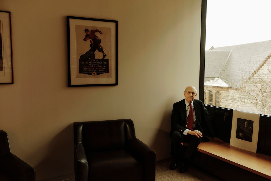 An older White man wearing glasses, a black suit, striped shirt and red tie, sits on a bench in the corner of his office. 