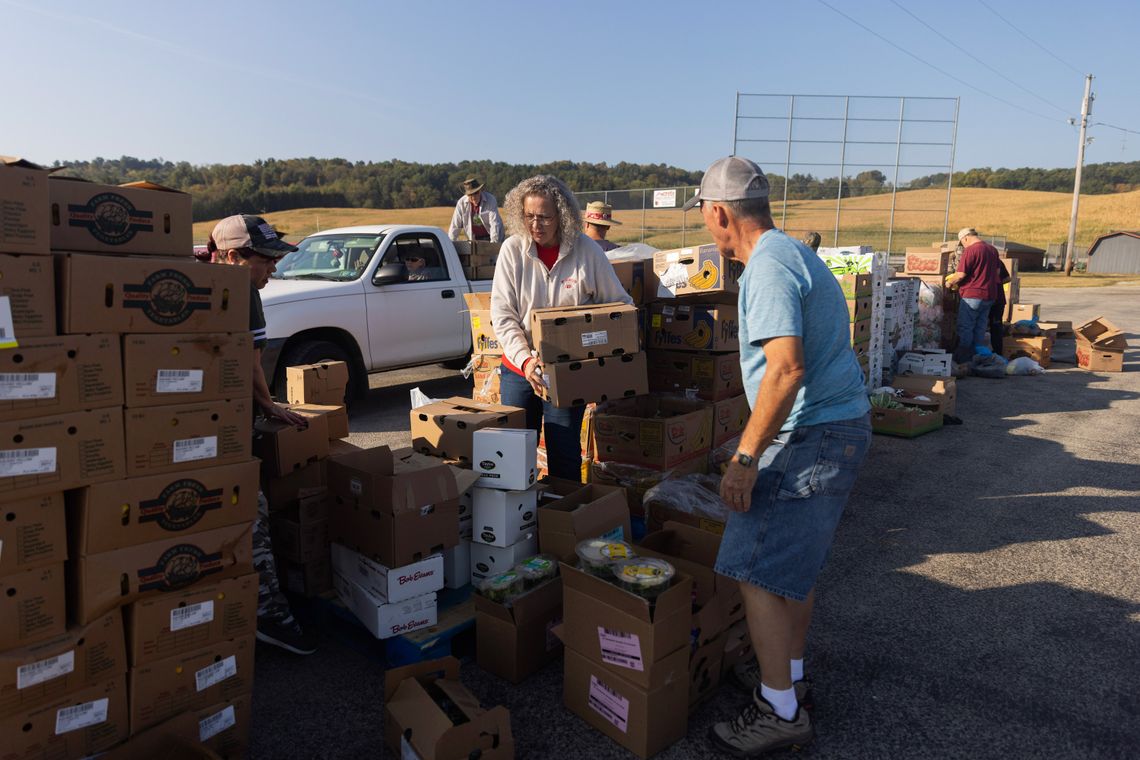 A photo of a White woman with curly gray hair lifting boxes. Stacks of cardboard boxes and other volunteers surround her. 