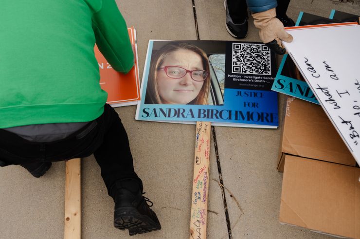 Signs lie on the ground as a person hovers over one on the left side of the photo, and another person holds one with a gloved hand.  A sign in the center reads: "Justice for Sandra Birchmore", and shows a picture of Sandra, a White young woman with brown hair and red-rimmed glasses.  