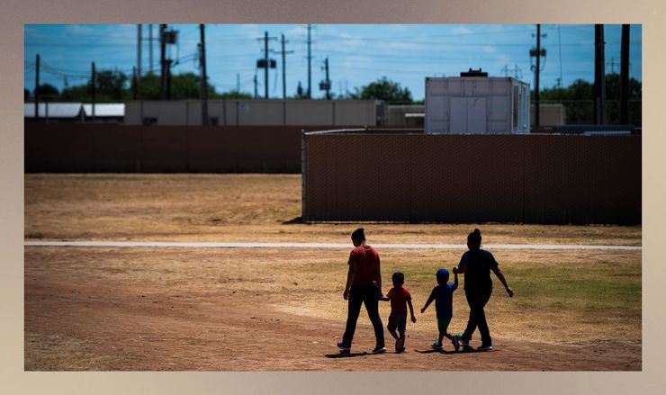 Two women and two children walk together outdoors with their backs facing the camera. Fencing is visible in front of them, along with some rectangular structures and power lines. The grass on the ground is dry.    