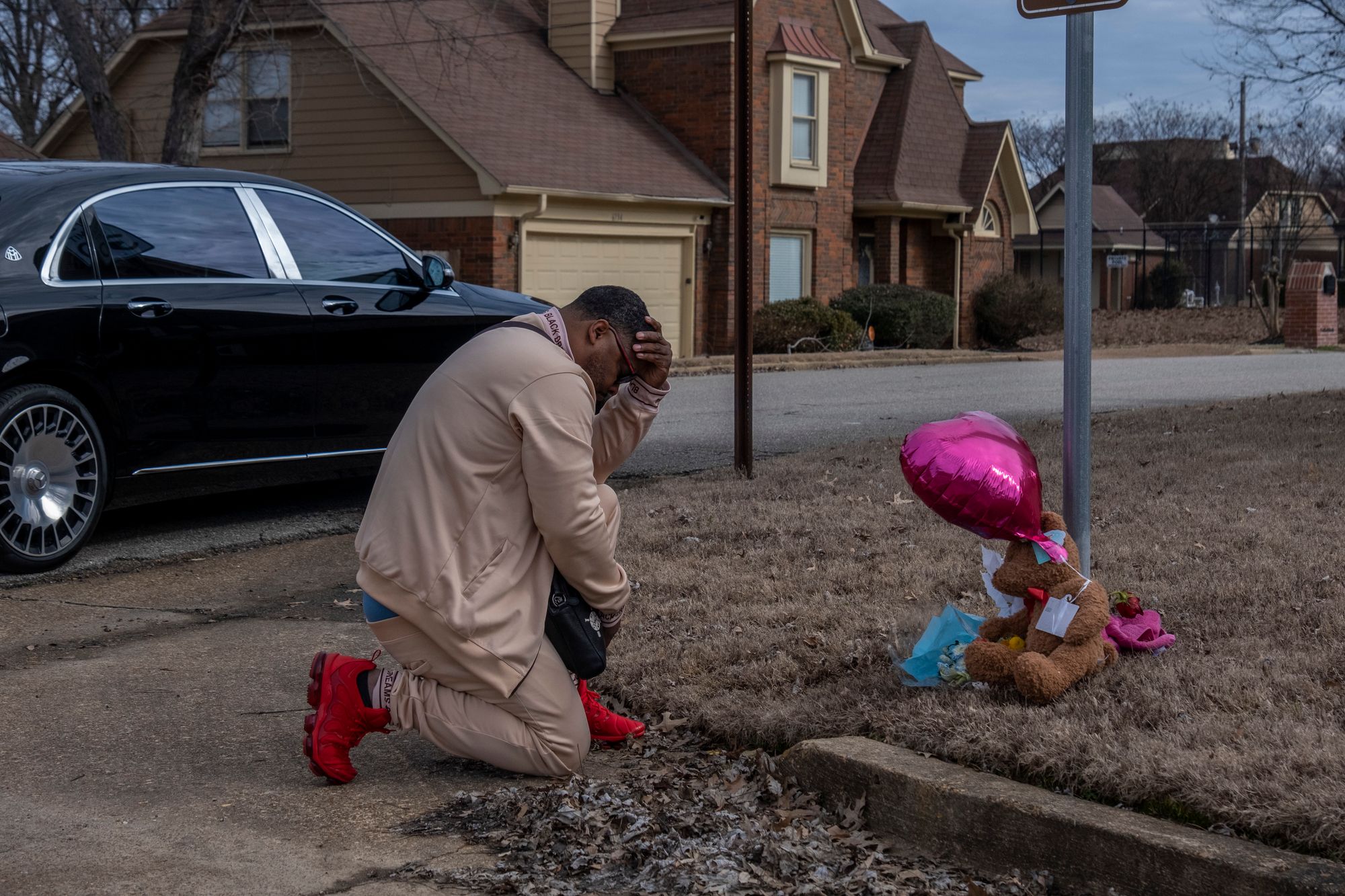 A Black man kneels at the side of a street, where a memorial is decorated with flowers, a teddy bear, and a pink balloon. 